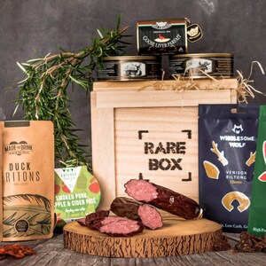 May include: A wooden crate with the words "Rare Box" printed on the front. The crate is filled with various gourmet food items, including chorizo thins, duck ritz, smoked pork apple and cider pate, venison biltong, and beef biltong.