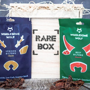 May include: Two packages of dried meat snacks in a wooden box. The box is labeled "Rare Box". The packages are labeled "Wholesome Wolf" and feature a deer and a bull. The deer package is blue with gold accents and says "Venison Biltong Classic". The bull package is green with red accents and says "Beef Biltong Moorish".