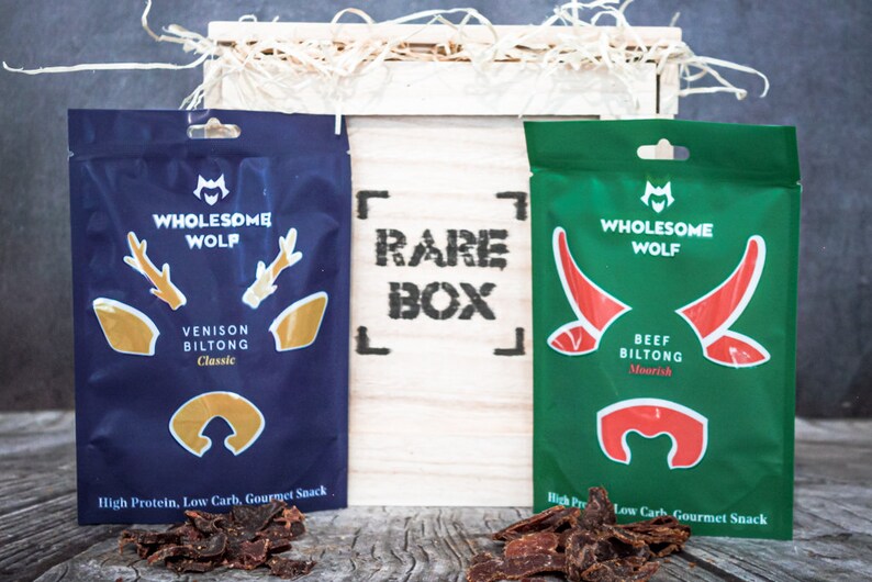 May include: Two packages of dried meat snacks in a wooden box. The box is labeled "Rare Box". The packages are labeled "Wholesome Wolf" and feature a deer and a bull. The deer package is blue with gold accents and says "Venison Biltong Classic". The bull package is green with red accents and says "Beef Biltong Moorish".