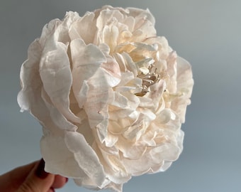 May include: A delicate, off-white peony flower with a slightly pink hue. The flower is made of paper and has a realistic appearance.