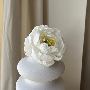 May include: A white ceramic vase with a textured finish, shaped like stacked circles. A single white peony with yellow center is in the vase.