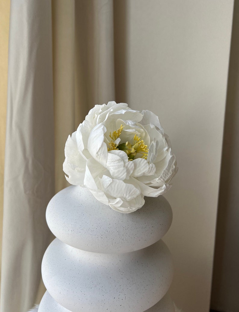 May include: A white ceramic vase with a textured finish, shaped like stacked circles. A single white peony with yellow center is in the vase.