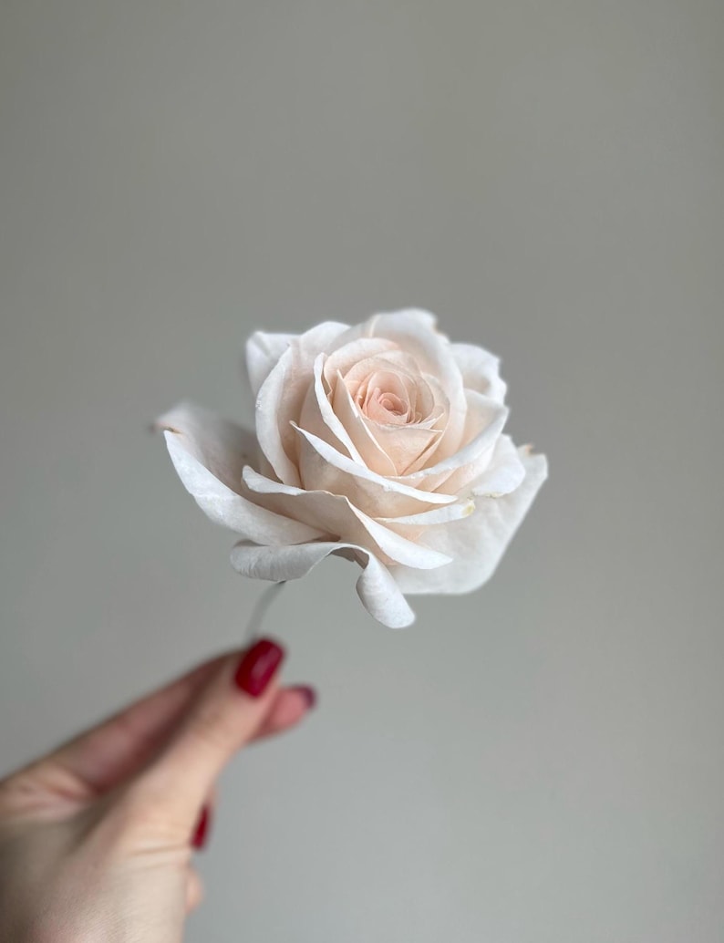 May include: A single artificial rose flower featuring delicate, layered petals in a soft, pale cream and blush pink color palette. The bloom is fully opened, showcasing a structured, realistic form with a thin, slender stem extending downward.