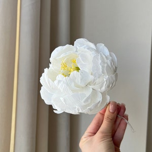 May include: A single white paper peony flower with a yellow center. The flower is held in a hand against a white background.
