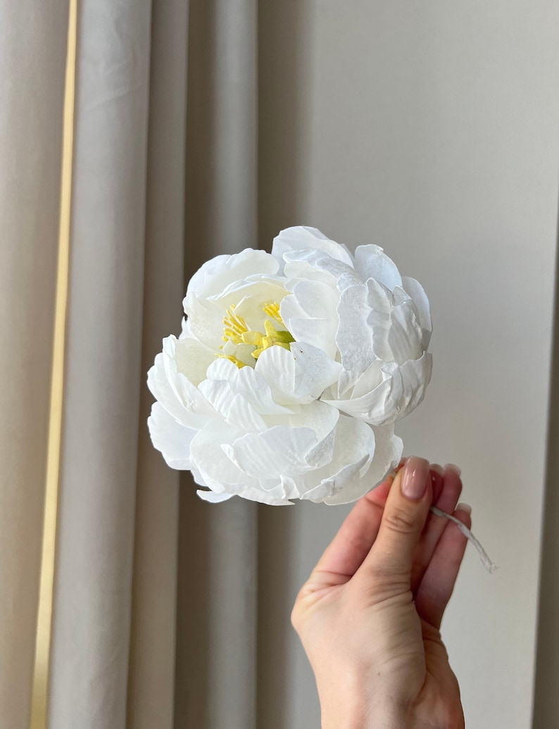 May include: A single white paper peony flower with a yellow center. The flower is held in a hand against a white background.