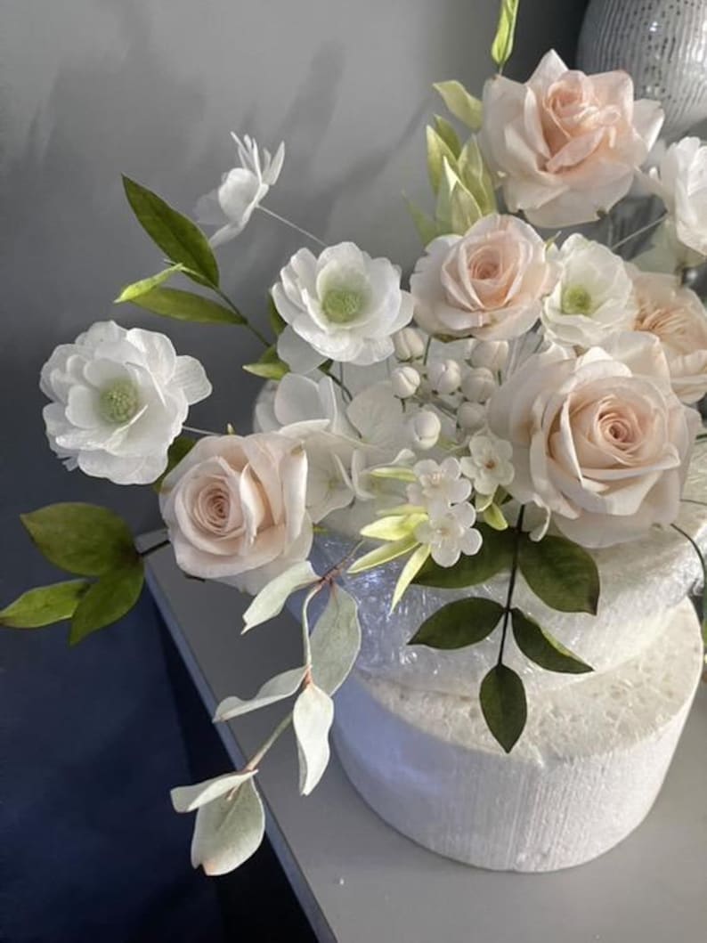 May include: A floral arrangement featuring pale pink and white roses, small white blossoms, and green leaves. The flowers are attached to a white, circular foam base wrapped in clear plastic, creating a decorative display of botanical elements.