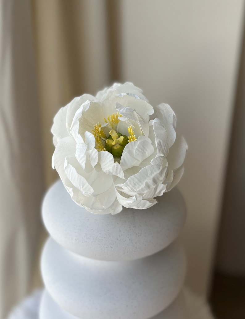 May include: A white ceramic vase with a stack of round, textured white ceramic pieces. A single white flower with a yellow center sits on top of the vase.