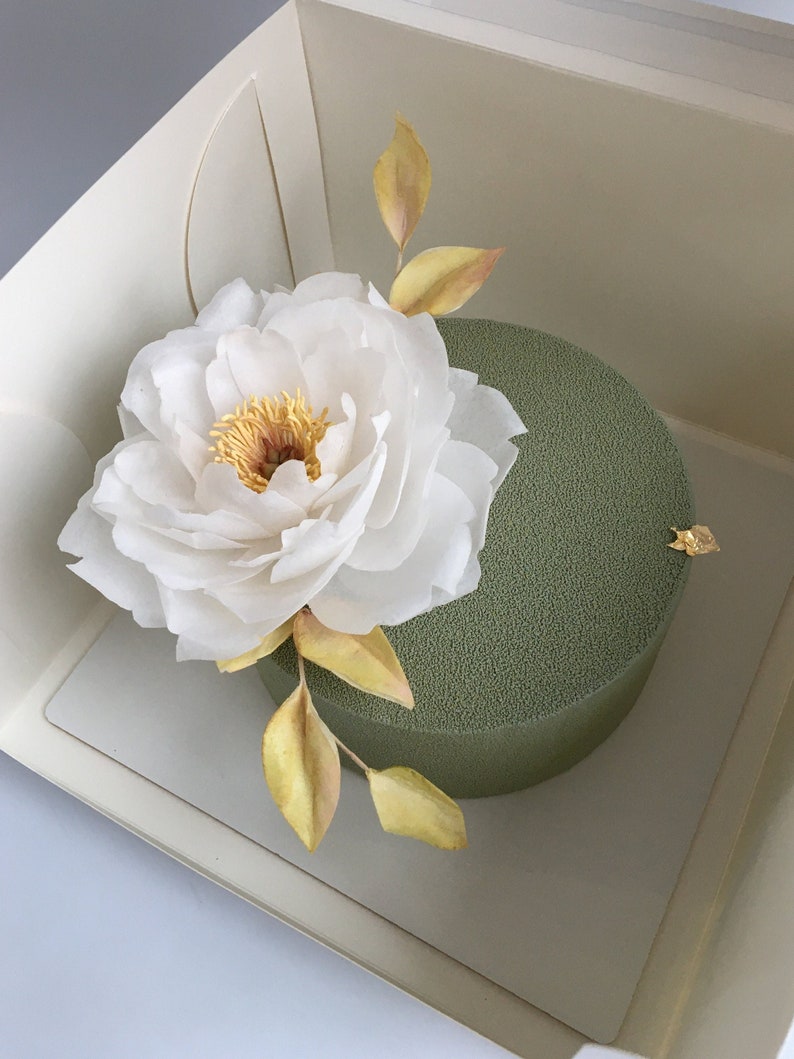 May include: A green cake with a white peony flower and gold leaves on top. The cake is in a white box.