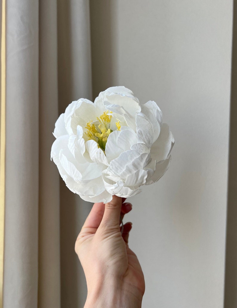 May include: A single white peony flower with yellow center. The flower is held in a hand.