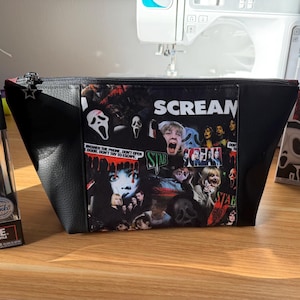 May include: A black zipped pouch featuring a collage of images from the Scream film franchise, including the Ghost Face mask and the text SCREAM. The pouch is flanked by two Funko Pop Ghost Face vinyl figures in their original display boxes.