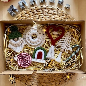 May include: A festive holiday-themed arrangement featuring a Santa Claus figurine, stone elephant figurines, and a box filled with macrame ornaments. The ornaments include a gnome, a wreath, a heart, a tree, a rainbow, and a candy cane.