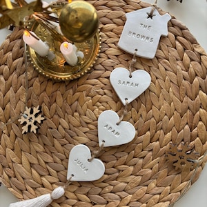 Puede incluir: Adorno de nombre familiar de cerámica blanca con los nombres "THE BROWNS", "SARAH", "ADAM" y "JULIA". El adorno está ensartado con hilo y lleva una borla. El fondo incluye un salvamanteles tejido, velas y copos de nieve decorativos.