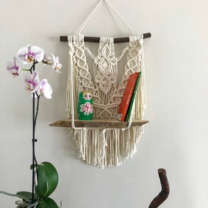 Puede incluir: Una estantería de pared de macramé con una base de madera y cordones de algodón blancos. La estantería está decorada con una muñeca rusa verde y roja y dos libros. Una planta de orquídea blanca está en primer plano.
