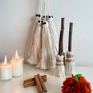 Könnte beinhalten: Ein Set aus fünf weißen Garn-Geister-Ornamenten mit schwarzen Augen, die an einer weißen Wand hängen. Zwei braune Reisigbesen mit weißem Garn um die Griffe und weißen Garnborsten stehen auf einer weißen Oberfläche. Auf der Oberfläche liegen drei Zimtstangen und zwei weiße Kerzen. Ein großer orangefarbener Garnkürbis liegt auf der Oberfläche.