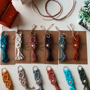 Könnte beinhalten: Sortiment an bunten Makramee-Schlüsselanhängern mit Holzperlen und Quasten. Jeder Schlüsselanhänger hat eine andere Farbe und ein anderes Muster. Die Schlüsselanhänger werden auf braunem Karton mit dem Text "Love because you loved me" präsentiert.