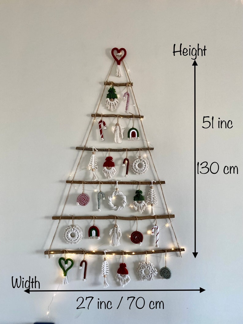 May include: A white wall with a macrame Christmas tree hanging on it. The tree is made of twine and has a variety of ornaments, including candy canes, stars, and hearts. The tree is 51 inches tall and 27 inches wide.