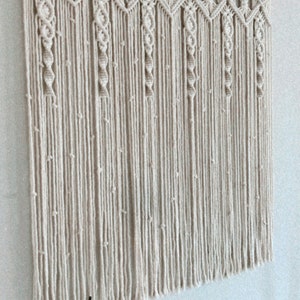 May include: A beige macrame wall hanging with a geometric pattern and fringe. The hanging is attached to a wooden dowel rod.