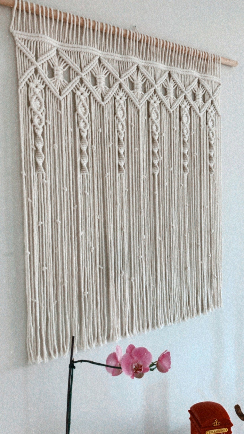 May include: A beige macrame wall hanging with a geometric pattern and fringe. The hanging is attached to a wooden dowel rod.