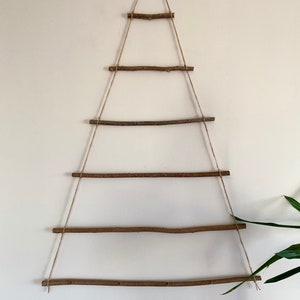 May include: A rustic Christmas tree made from branches and twine, hanging on a white wall. The tree is decorated with five horizontal branches, each tied to the twine with knots.