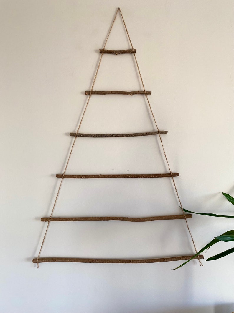 May include: A rustic Christmas tree made from branches and twine, hanging on a white wall. The tree is decorated with five horizontal branches, each tied to the twine with knots.