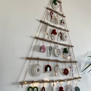 May include: A white wall with a Christmas tree made of branches and twine. The tree is decorated with handmade ornaments, including hearts, candy canes, gnomes, and rainbows. The ornaments are made of yarn and fabric in red, green, white, and brown.