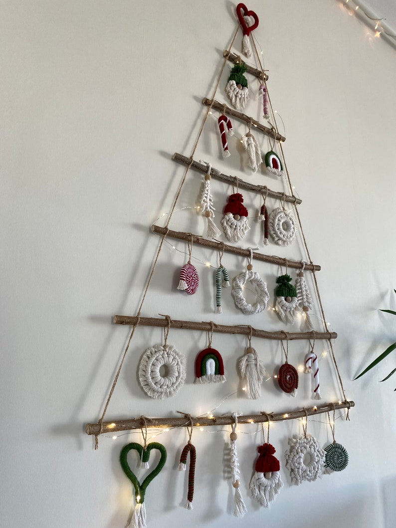 May include: A white wall with a Christmas tree made of branches and twine. The tree is decorated with handmade ornaments, including hearts, candy canes, gnomes, and rainbows. The ornaments are made of yarn and fabric in red, green, white, and brown.
