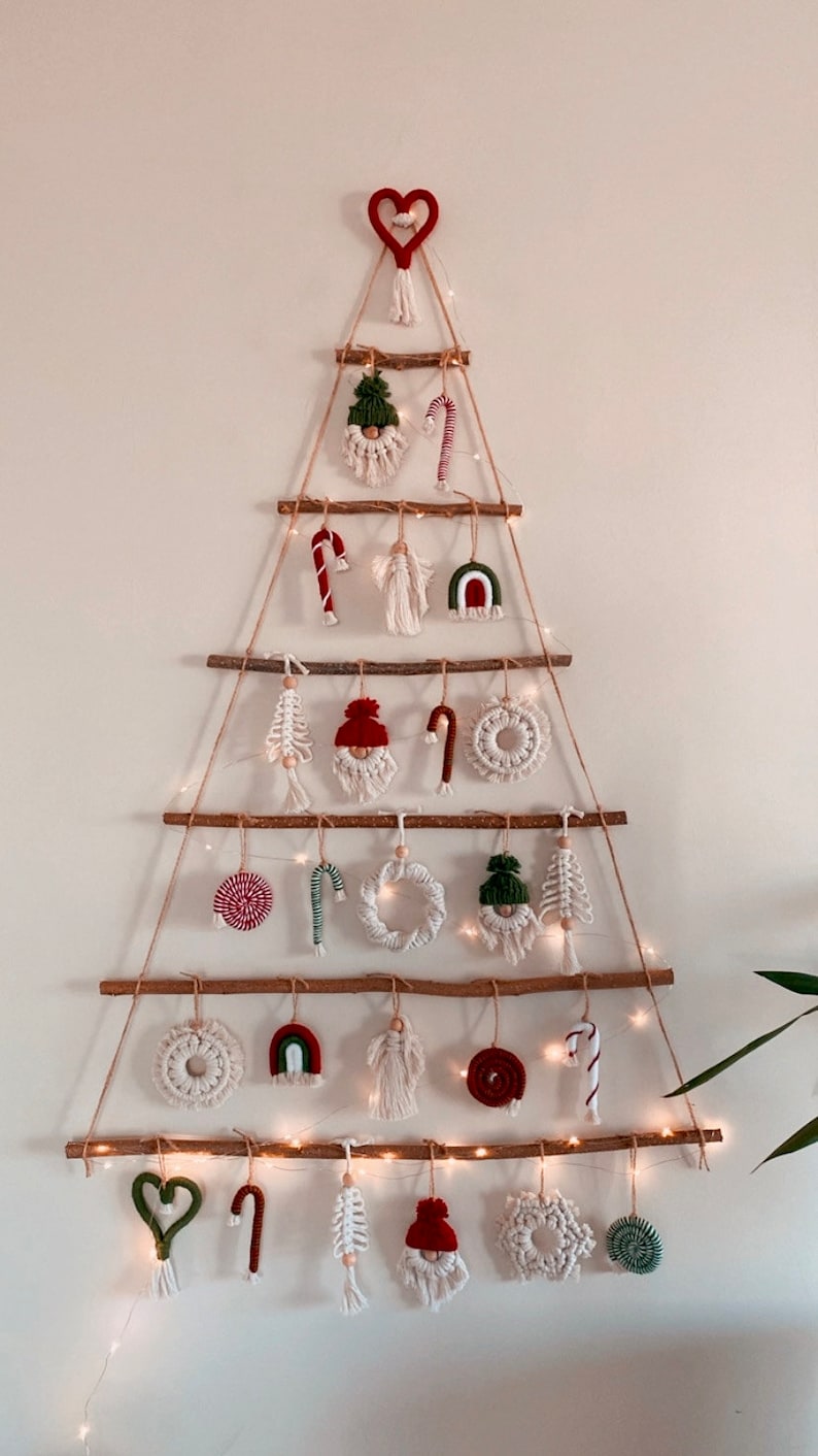May include: A white wall with a Christmas tree made of branches and string. The tree is decorated with handmade ornaments, including gnomes, candy canes, hearts, and rainbows. The ornaments are made of yarn and fabric.