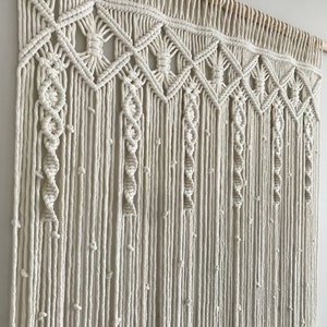 May include: A white macrame wall hanging with a geometric pattern. The hanging is made of cotton cord and has a wooden dowel at the top.