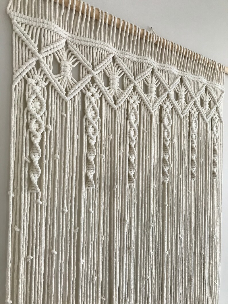 May include: A white macrame wall hanging with a geometric pattern. The hanging is made of cotton cord and has a wooden dowel at the top.