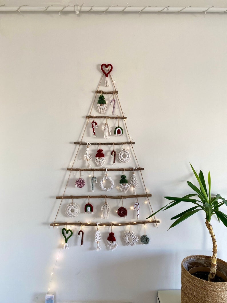 May include: A white wall with a Christmas tree made of branches and twine. The tree is decorated with red and green ornaments, including candy canes, hearts, and a small gnome. A string of white lights is strung around the tree. A potted plant is on the right side of the image.