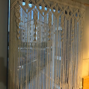 May include: A white macrame curtain with a geometric pattern hangs in a window. The curtain is made of thick cotton cord and has a fringe at the bottom.