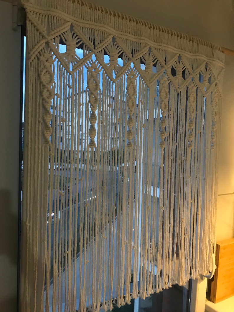 May include: A white macrame curtain with a geometric pattern hangs in a window. The curtain is made of thick cotton cord and has a fringe at the bottom.