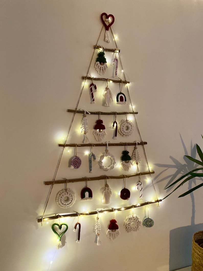May include: A white wall with a Christmas tree made of branches and string lights. The tree is decorated with handmade ornaments, including a red heart, a candy cane, a gnome, and a wreath.