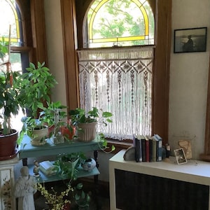 May include: A white macrame curtain hangs in a window with a stained glass design. The curtain is made of knotted cords and has a geometric pattern. There are plants in pots on a table in front of the window.