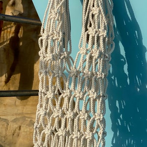 May include: A white crocheted tote bag hanging on a blue surface. The bag is made of thick rope and has a large, open design.