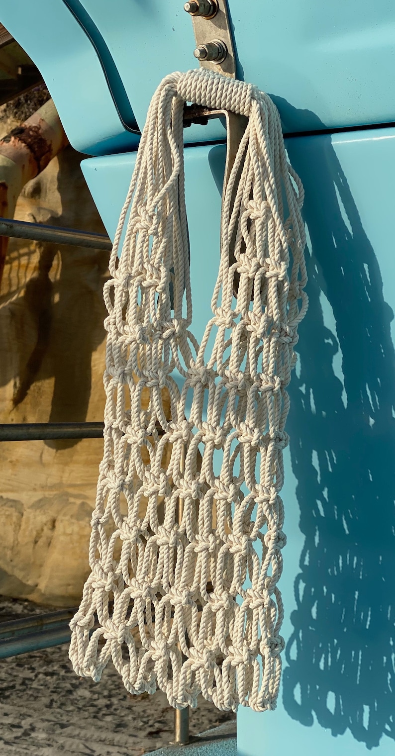 May include: A white crocheted tote bag hanging on a blue surface. The bag is made of thick rope and has a large, open design.