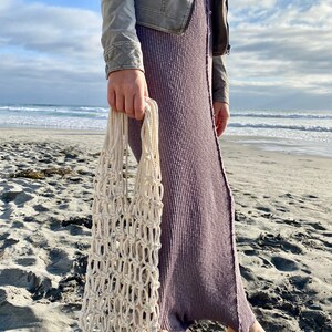 May include: A person is holding a white crocheted mesh bag on a sandy beach. The bag is made of thick rope and has a large opening at the top.