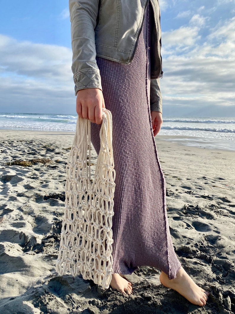 May include: A person is holding a white crocheted mesh bag on a sandy beach. The bag is made of thick rope and has a large opening at the top.