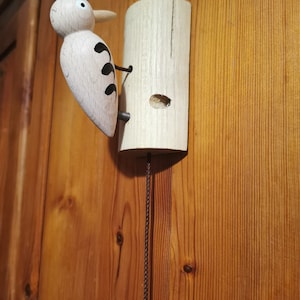 May include: Wooden woodpecker bird figurine with a brown string attached to a white ball. The bird is perched on a wooden tree trunk with a hole in it. The background is a wooden wall.