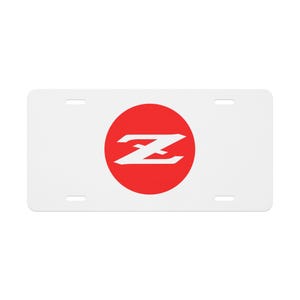 May include: White license plate featuring a red circle with a white stylized "Z" logo. The plate has four mounting holes and a rectangular shape. The design is simple and bold.