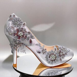 Könnte beinhalten: Ein silberner High-Heel-Pumps aus Satin mit aufwendiger Blumenstickerei aus Silberfaden, klaren Pailletten und kleinen weißen Perlen. Die Verzierungen konzentrieren sich auf den Zehen- und Fersenbereich und erzeugen ein strukturiertes Aussehen.