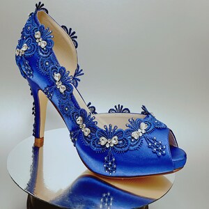May include: Royal blue satin high heel shoes with a peep toe and intricate lace detailing. The shoes are adorned with rhinestones and have a delicate floral design.