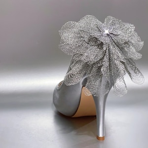 Könnte beinhalten: Ein silberner High-Heel-Schuh mit glatter Satin-Oberfläche an Absatz und Rückseite. Eine große, dekorative, gerüschte Blume aus metallischem silbernem Netzstoff ist am Absatz befestigt und mit einem kleinen, klaren, funkelnden Kristall verziert.