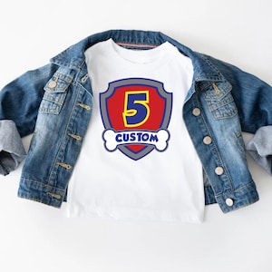 Op de afbeelding: Wit T-shirt met een kleurrijke afbeelding met het nummer 5 in geel, een rood schild en het woord "CUSTOM". Het shirt wordt getoond op een spijkerjasje.