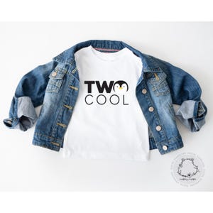 May include: White toddler t-shirt with the text "TWO COOL" and a penguin graphic. The shirt is paired with a blue denim jacket. The image is set against a white background, creating a clean and simple aesthetic.