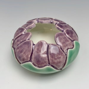 May include: A ceramic bowl with a floral design. The bowl features a ring of purple petal-like shapes around the rim, with a light green base. The interior of the bowl is a light cream color.