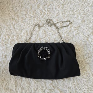 May include: Black clutch purse with a silver chain strap. The purse has a gathered design and a decorative embellishment in the center. The background is a white, textured surface.