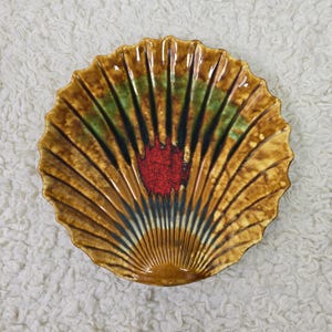 Puede incluir: Un plato decorativo de cerámica con forma de concha. El plato presenta una combinación de colores marrón, verde y rojo con un diseño en forma de abanico. El plato tiene un borde festoneado.