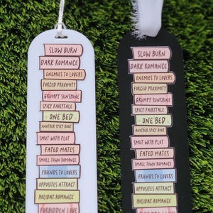May include: Two bookmark designs with a stack of books and different romance tropes. The bookmarks are white and black with pink, yellow, and blue text. The text on the bookmarks reads: "Slow Burn Dark Romance", "Enemies to Lovers", "Forced Proximity", "Grumpy Sunshine", "Spicy Fairytale", "One Bed", "Another Spicy Book", "Smut with Plot", "Fated Mates", "Small Town Romance", "Friends to Lovers", "Opposites Attract", "Holiday Romance", and "Forbidden Love".