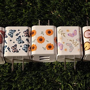 May include: Five white phone chargers with different colorful patterns. The patterns include hearts, butterflies, sunflowers, and a smiley face flower.